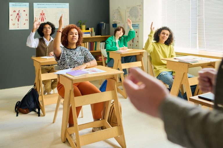 Group of intercultural teenage students of high school raising hands in a classroom
